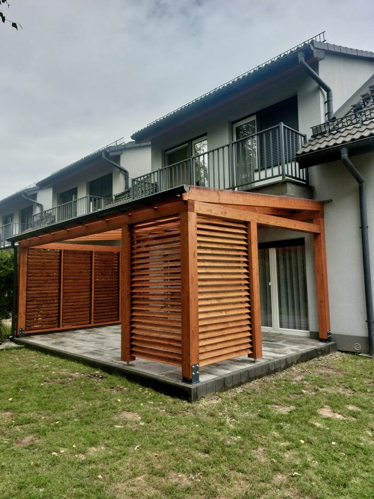 Zdjęcie przestawia realizację zadaszenia tarasu wykonanego z drewna klejonego BSH, pokryty poliwęglanem litym, wykończenie ścian w systemie ruchomych deseczek